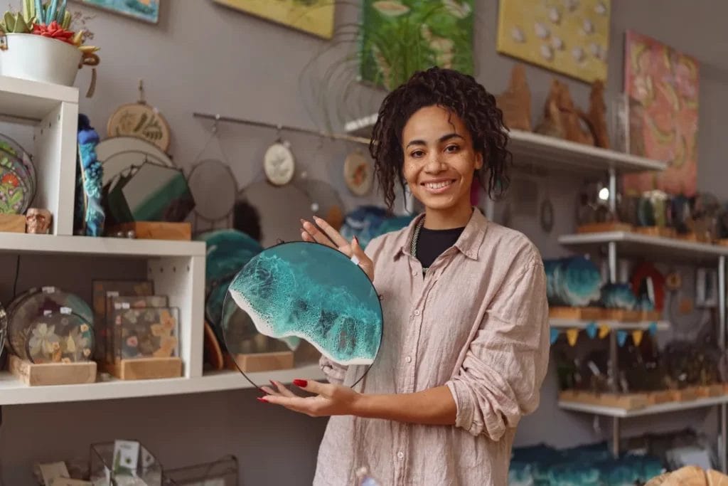An artist wearing a light beige shirt holds a round glass and resin piece depicting ocean waves in an artist's studio.