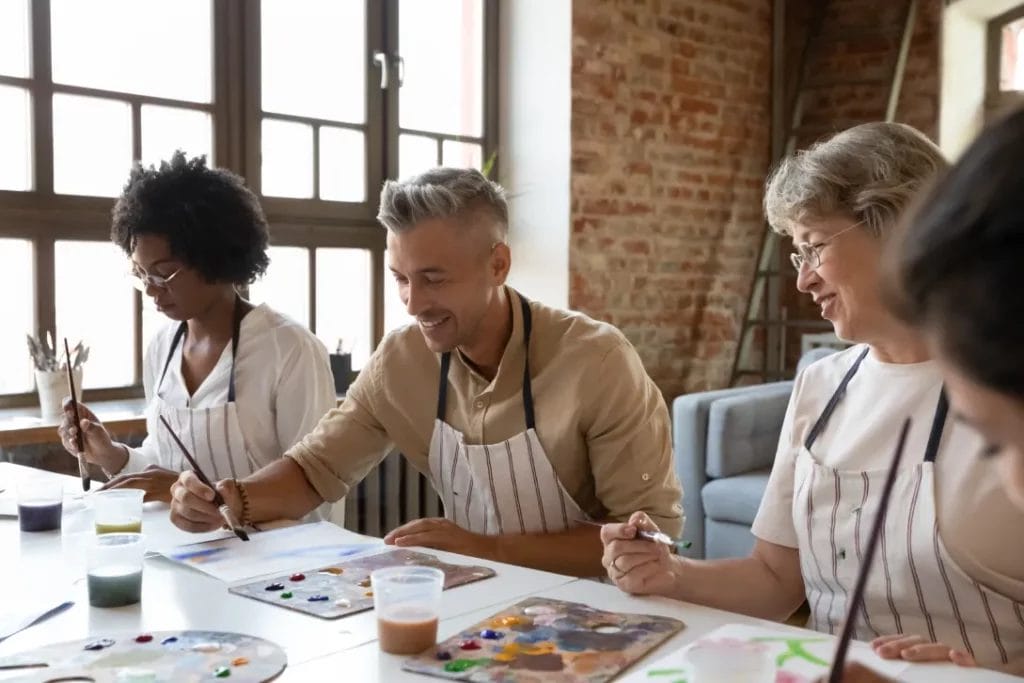 A group of people wearing white striped aprons sit at a white table and paint during an art class.