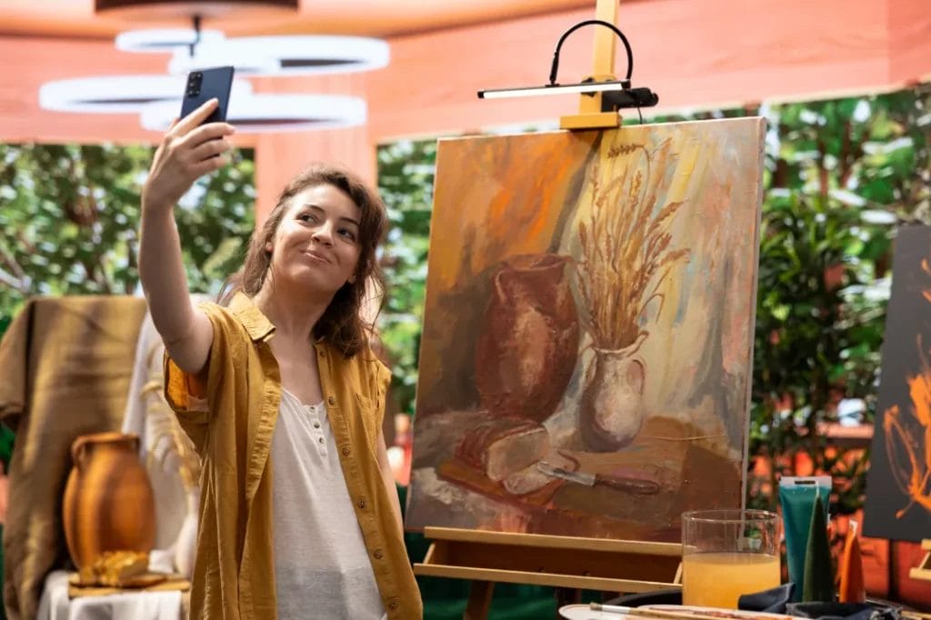 An artist wearing a mustard shirt smiles and takes a selfie with her canvas, depicting a still life of a pitcher, vase, and bread in neutral tones, in the background.