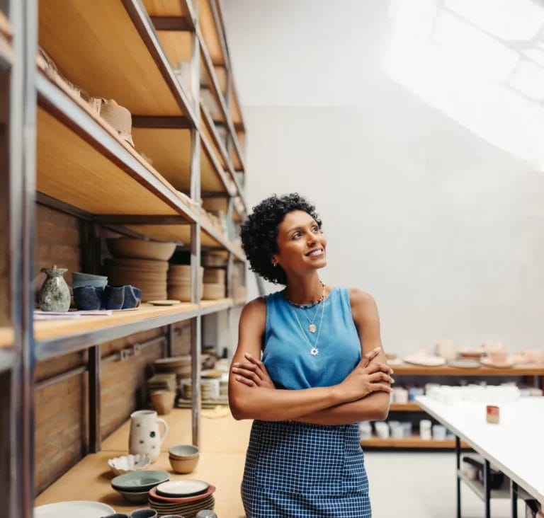 An artist wearing a blue tank top and blue plaid skirt folds her arms, smiles, and looks up to the side in a pottery studio with art pieces on shelves next to her.
