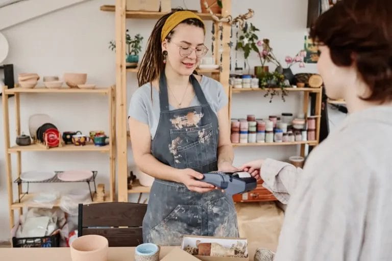 Young female owner of small pottery shop with payment terminal selling handmade items to client paying for purchase by credit card