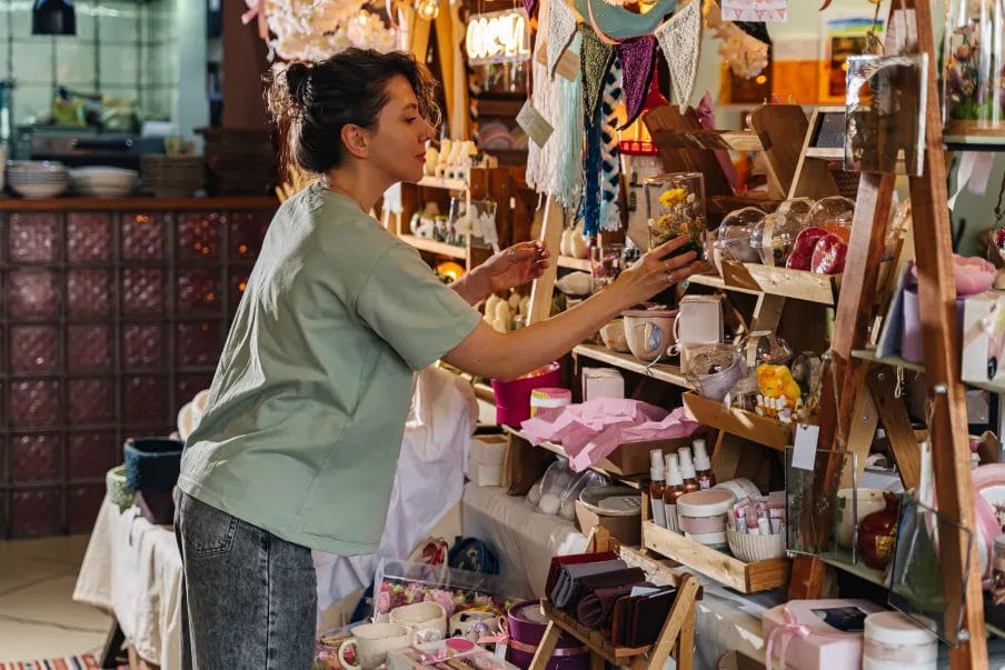 An artist wearing a mint top and blue jeans arranges products on the shelves of her vendor booth display.