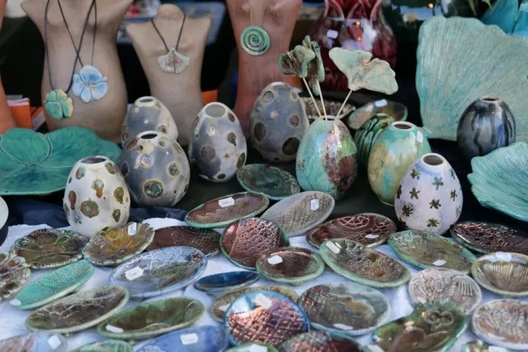 Ceramic dishes, vases, and necklaces in tones of blue, gray, and teal are arranged on an art vendor's booth table.