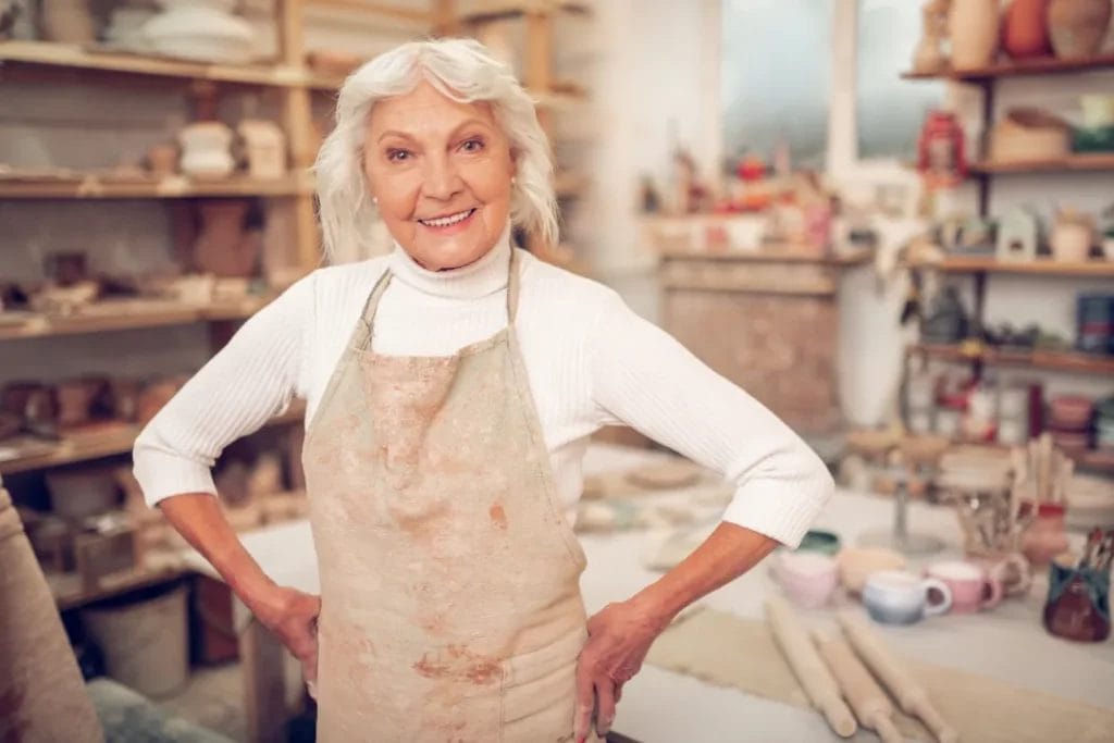A woman with white hair wearing a white long-sleeve top and taupe apron smiles in her ceramic studios.