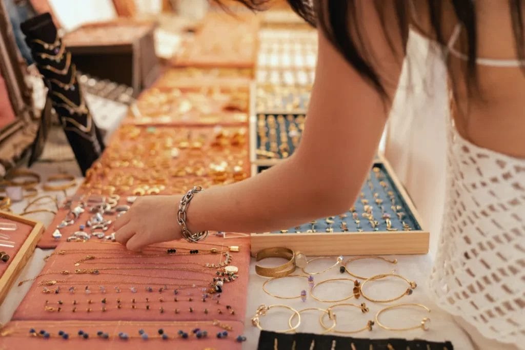 A market attendee shops for jewelry at a jewelry maker's stand, featuring necklaces, goldtone bangles and rings on red and jewelry box displays.