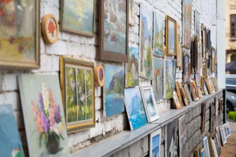 An image of nature paintings in various frames against a white brick wall at an art fair.