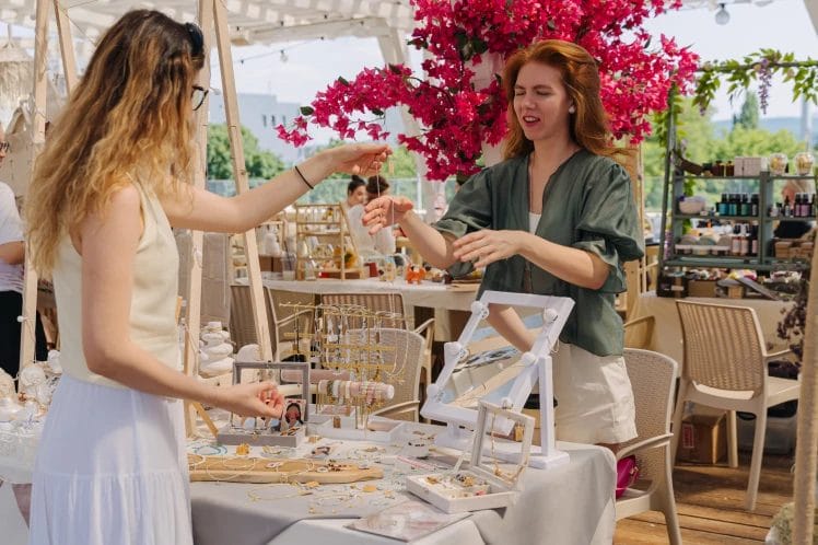 handmade jewelry seller demonstrating accessories to a customer at an outdoor craft market