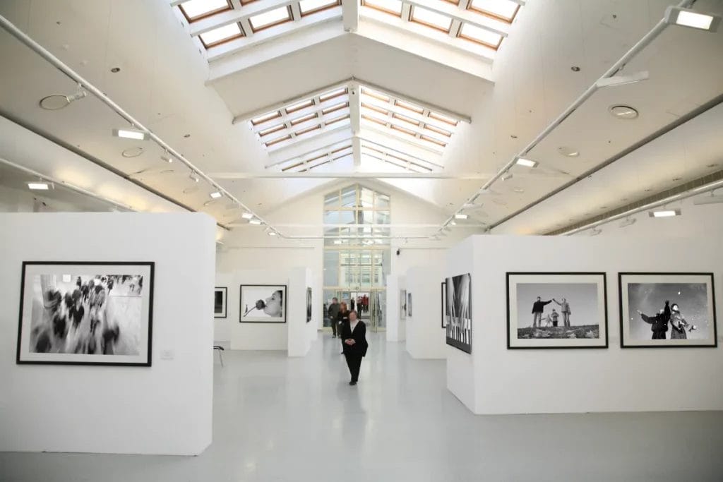An art expo displaying black-and-white photographs on white walls in a large white room with ceiling windows and lighting fixtures.