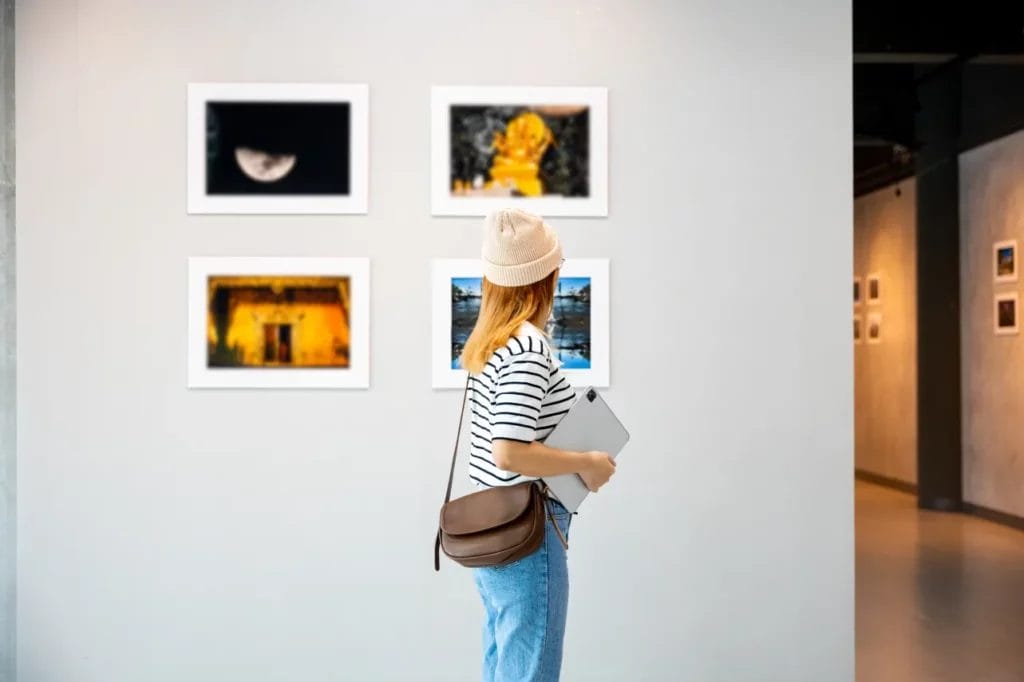 An artist wearing a black-and-white striped tee, blue jeans, and a cream-colored beanie holds a laptop and looks at a display of photographs on a white wall in an art gallery.