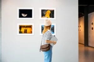 An artist wearing a black-and-white striped tee, blue jeans, and a cream-colored beanie holds a laptop and looks at a display of photographs on a white wall in an art gallery.