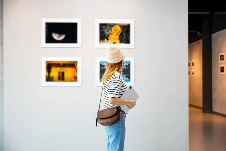 An artist wearing a black-and-white striped tee, blue jeans, and a cream-colored beanie holds a laptop and looks at a display of photographs on a white wall in an art gallery.