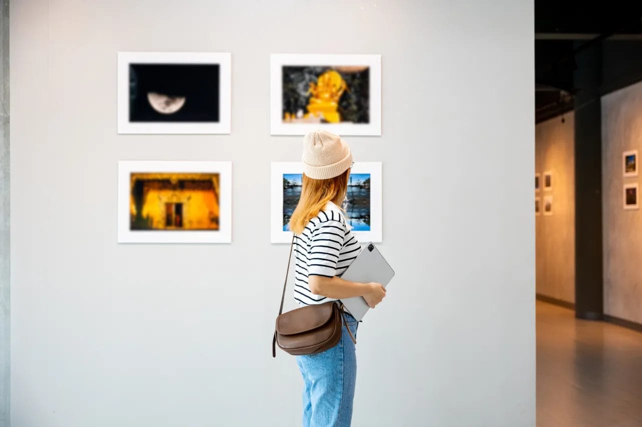 An artist wearing a black-and-white striped tee, blue jeans, and a cream-colored beanie holds a laptop and looks at a display of photographs on a white wall in an art gallery.