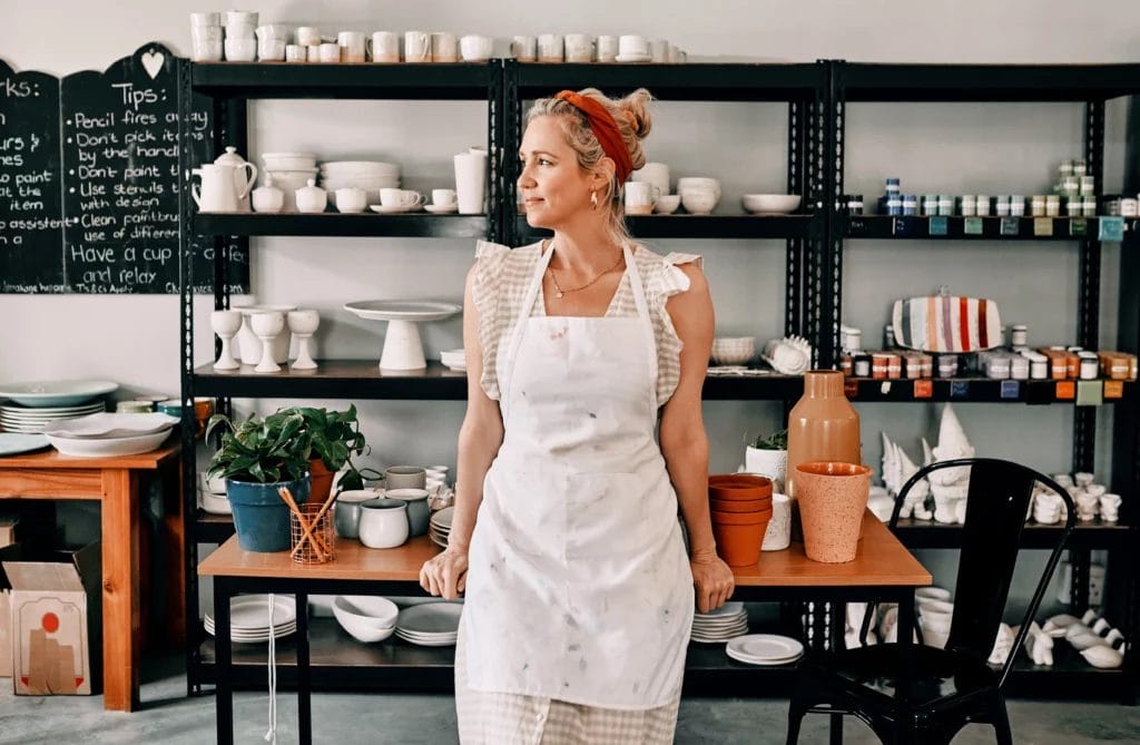 woman potter standing in a studio workshop with a shelf full of ceramics and glazes
