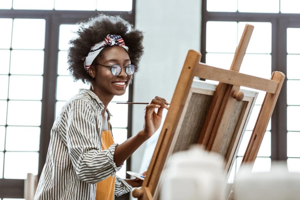 An artist wearing a patterned headscarf, a striped shirt, and an orange blouse smiles while painting on a canvas in a studio with large windows in the background.