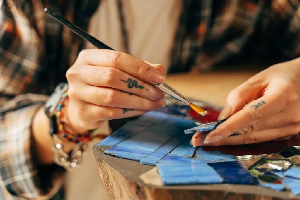 A close-up image of an artist's hands painting blue tiles for a mosaic piece.