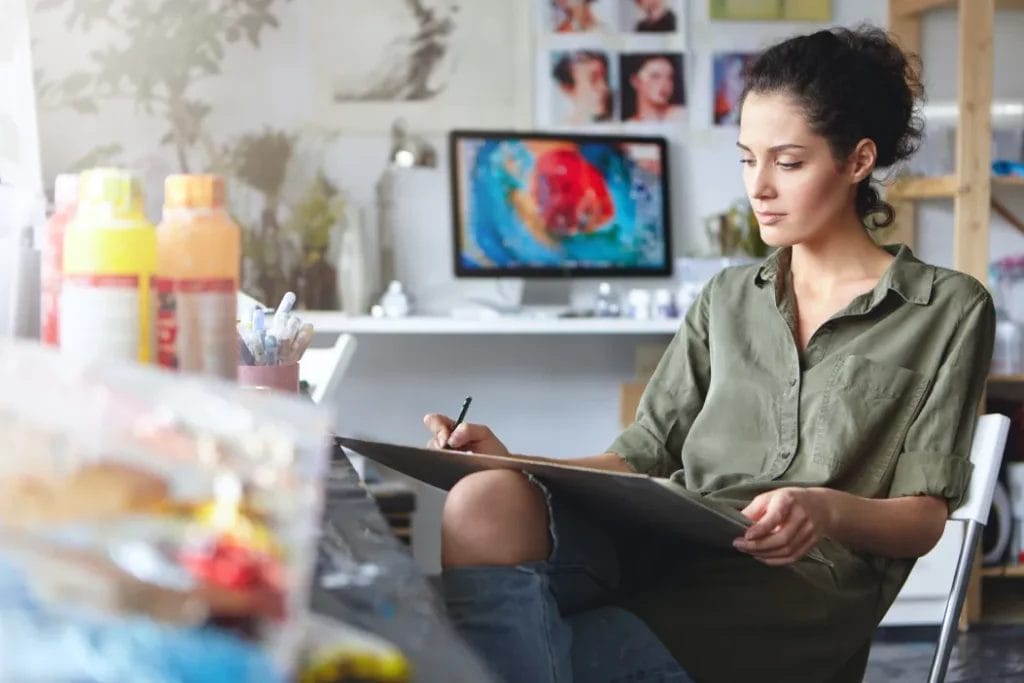 An artist sits and writes notes on a large writing pad in an art studio with various art supplies set on a table in the foreground.