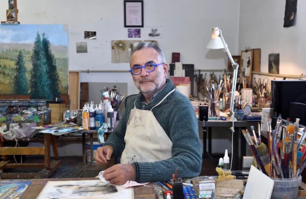 An artist wearing a green sweater and beige apron sits at a wooden desk in his art studio with a canvas depicting green trees, plus various paintbrushes and art tools in the background.