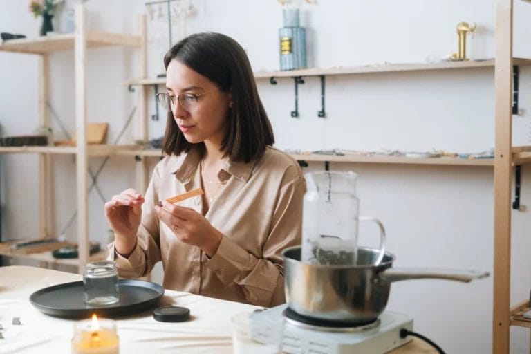 a woman sitting at a table using a wick and melted wax to make a handmade candle